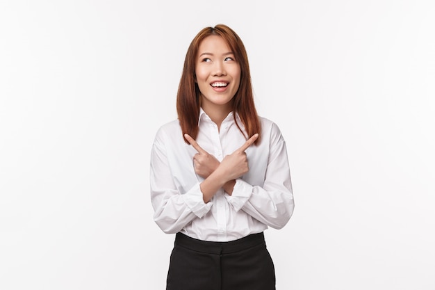 Retrato de divertida e sonhadora mulher asiática sorridente na camisa branca, desviar o olhar pensativo como fazer escolha, apontando para o lado esquerdo e direito, tem poucas variantes, em pé parede branca