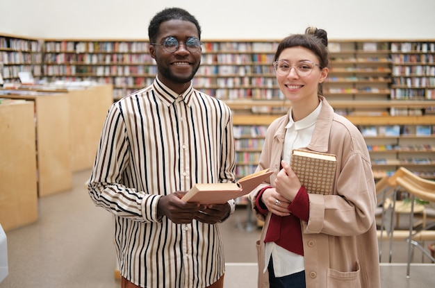 Retrato de dois alunos segurando livros e sorrindo para a câmera em pé na biblioteca