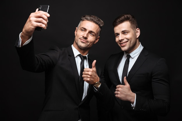 Retrato de dois empresários satisfeitos vestidos com um terno formal tirando foto de selfie no celular isolada sobre a parede preta