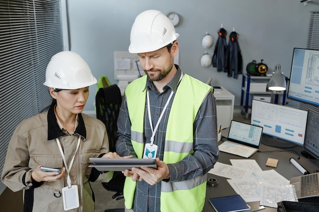 Retrato de dois engenheiros vestindo capacetes em ambientes fechados e revisando planos para copiar o espaço