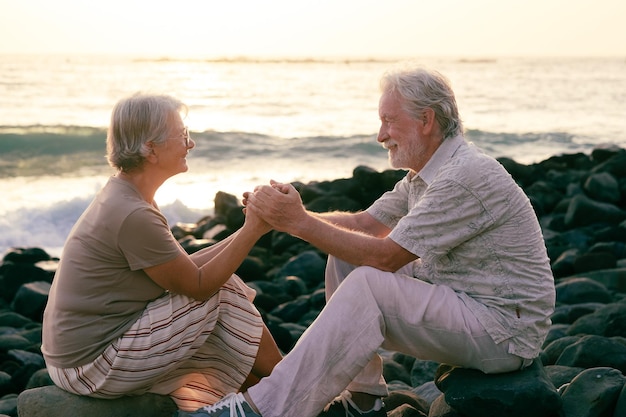 Retrato de dois idosos felizes e românticos ou pensionistas sentados na praia à luz do sol mãos nas mãos velho casal sênior sorridente ao ar livre curtindo férias juntos