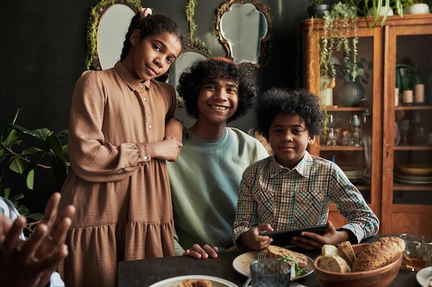 Retrato de dois irmãos e irmã sorrindo para a câmera enquanto está sentado à mesa de jantar durante a família di