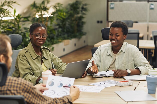 Retrato de dois jovens afro-americanos sorrindo para os colegas enquanto está sentado à mesa durante uma reunião em um escritório moderno e discutindo o projeto de trabalho