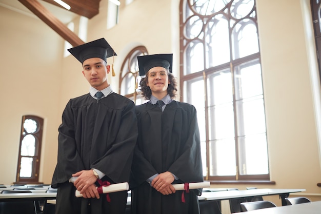 Retrato de dois jovens vestindo túnicas de formatura e olhando para a câmera enquanto está no auditório da escola, copie o espaço da cintura para cima