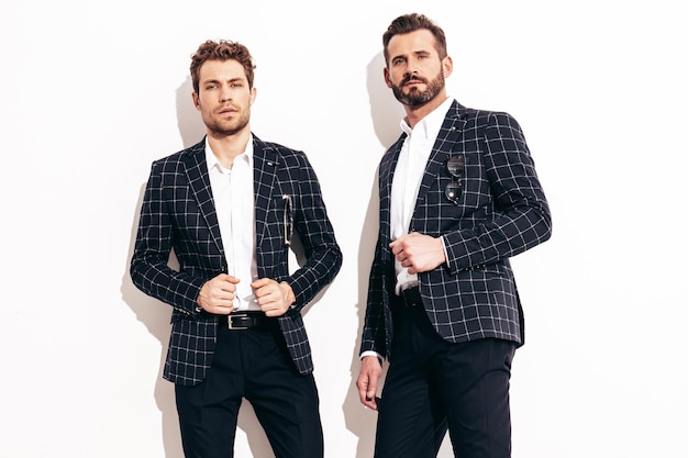 Retrato de dois modelos lambersexual hipster elegante e confiante bonito homens modernos sexy vestidos de terno preto elegante moda masculina posando no estúdio perto da parede branca