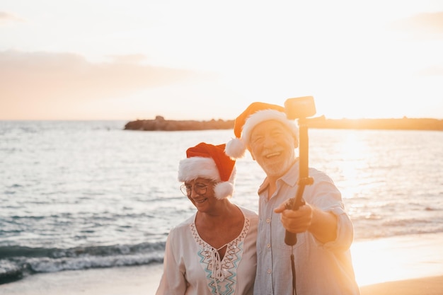 Retrato de dois velhos fofos se divertindo e curtindo juntos na praia nos dias de natal na praia usando chapéus de natal olhando e segurando uma câmera gravando vídeos de férias