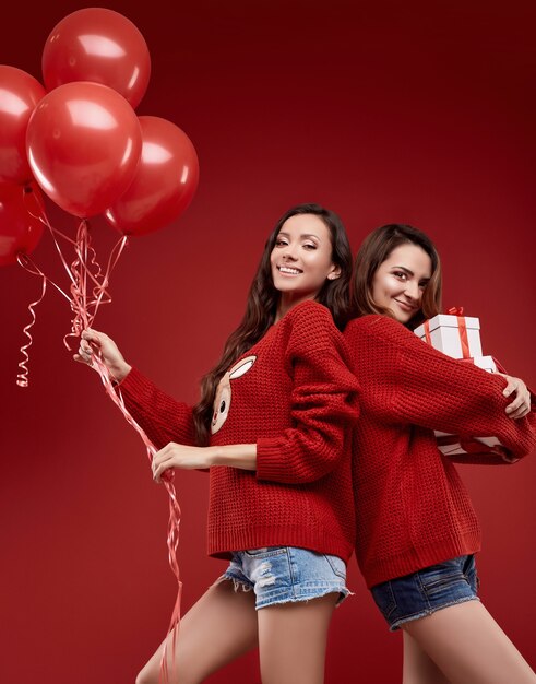 Foto retrato de duas amigas gêmeas malucas em um agasalho de inverno aconchegante com balões de festa e caixas de presente posando