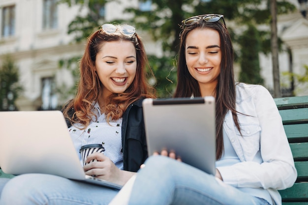 Retrato de duas belas mulheres olhando para um tablet rindo enquanto outra ruiva está segurando um laptop nas pernas e uma xícara de café enquanto está sentado em um banco.