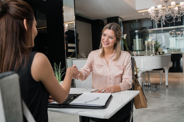 Retrato de duas jovens empresárias tendo reunião e aperto de mãos no saguão do hotel. Conceito de viagens de negócios.