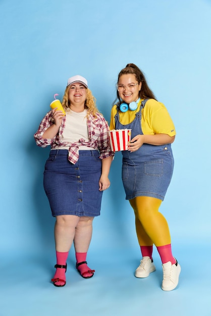 Retrato de duas lindas amigas gordas posando com cesta de pipoca sobre o fundo azul do estúdio Tempo de lazer de fim de semana