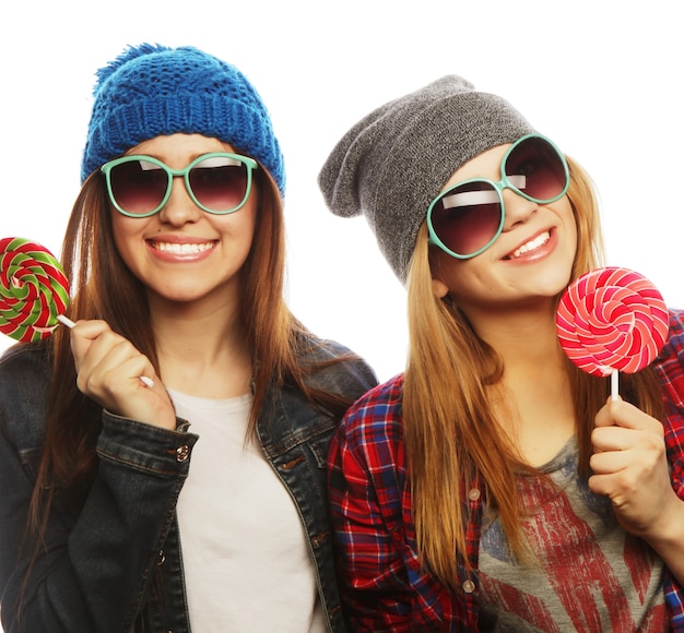 Retrato de duas meninas jovens bonitas hippie com chapéus e óculos de sol segurando doces. Retrato de estúdio de dois melhores amigos alegres, se divertindo e fazendo caretas.