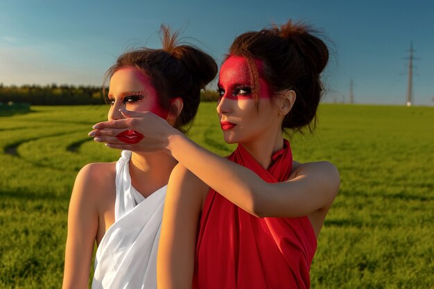 Retrato de duas meninas morenas com maquiagem criativa de um campo verde e um céu azul