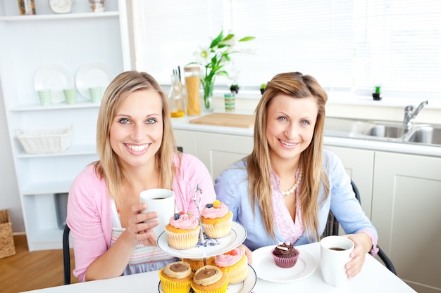 Retrato de duas mulheres atraentes com xícaras de café e cupcakes sentados na cozinha