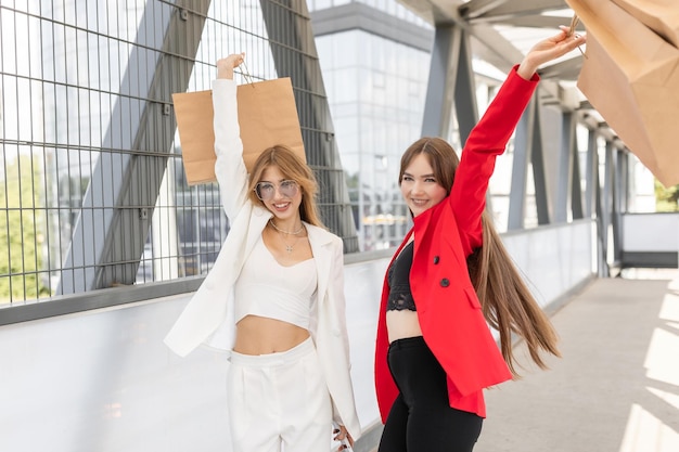 Retrato de duas mulheres felizes vestidas com ternos elegantes estão andando juntas perto do shopping com um monte de sacos de papel conceito de compras