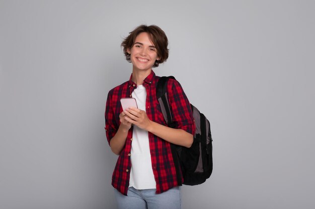 Retrato de elegante moderna sorridente jovem estudante com smartphone vestindo camisa a quadros em fundo cinza conversando surfing na internet online estudo em aplicativos de rede