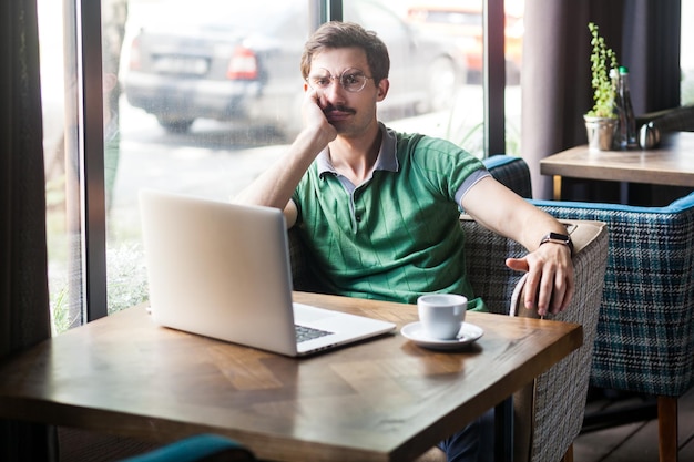 Retrato de empregado homem vestindo camiseta verde olhando para a câmera com expressão séria, mantendo a mão sob o queixo trabalhando on-line no laptop Tiro interno perto do fundo do café da janela grande