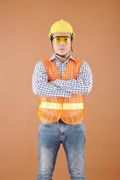 Retrato de empreiteiro confiante em colete brilhante, óculos de proteção e capacete de segurança, cruzando os braços e olhando para a câmera