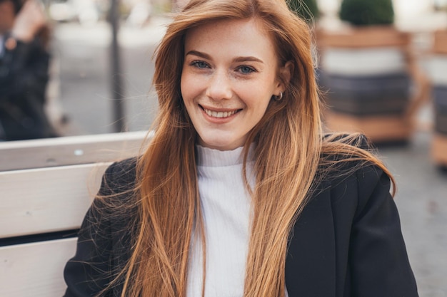 Foto retrato de empresária com cabelo longo e sexy e rosto sardento, olhando sorrindo para a câmera, sentado no banco em frente ao prédio de escritórios pessoa positiva retrato de negócios rosto feliz