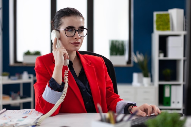 Retrato de empresária com óculos em telefonema sentado na mesa de inicialização. empresário de jaqueta vermelha ouvindo funcionário ao telefone. inicie o proprietário da empresa em uma conversa casual.