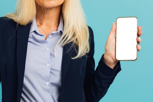 Retrato de empresária madura em traje formal segurando smartphone com tela de dispositivo em branco branco na mão perto da câmera, estúdio azul. Gadget com espaço livre para maquete