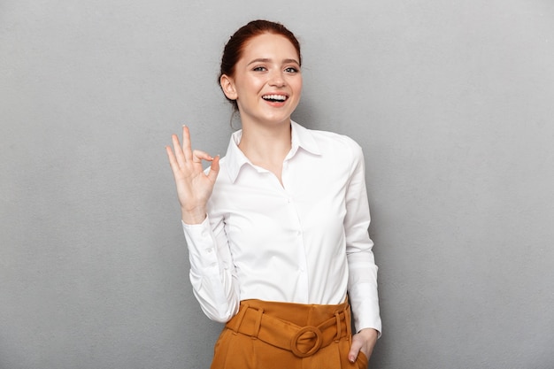 Retrato de empresária ruiva confiante de 20 anos em roupa formal sorrindo e mostrando sinal de ok no escritório isolado sobre o cinza