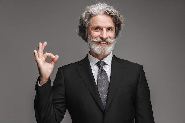 Retrato de empresário adulto alegre vestindo terno preto formal, sorrindo para a câmera e mostrando sinal de ok isolado sobre a parede cinza