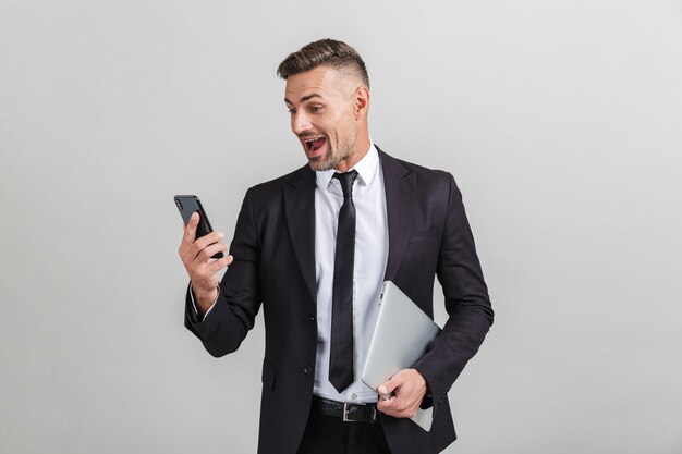 Retrato de empresário adulto animado em terno de escritório, olhando para o smartphone e segurando o laptop em pé isolado