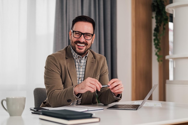 Retrato de empresário alegre com laptop e diário sentado na mesa do escritório