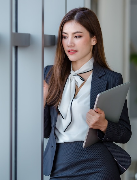 Retrato de empresário feminino asiático positivo ou secretário em roupas formais, em pé com o computador laptop netbook na sala do escritório e olhando para o ponto distante na pose de relaxamento.