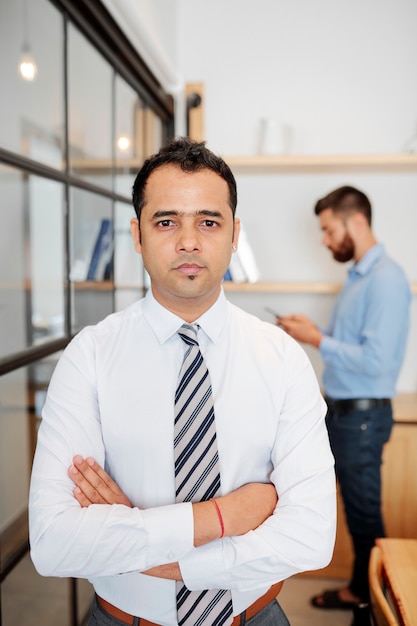 Retrato de empresário indiano sério e confiante, cruzando os braços e olhando para a câmera quando está no escritório