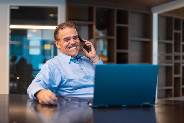 Retrato de empresário maduro usando laptop no escritório e falando no celular