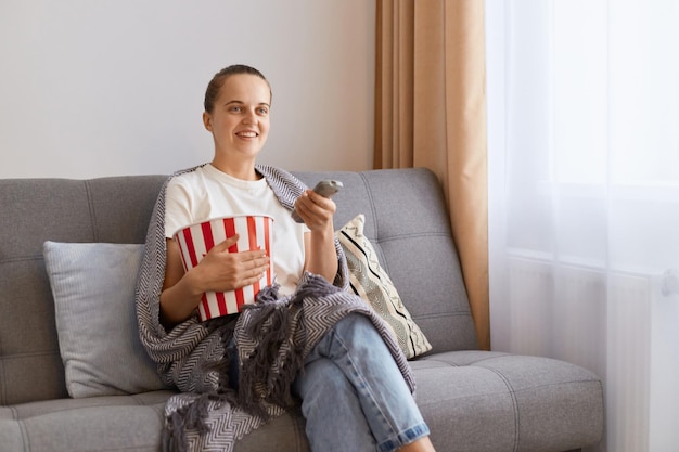 Retrato de encantadora jovem caucasiana adulta tendo tempo livre sentado no sofá assistindo séries engraçadas comendo pipoca sorrindo desfrutando de um filme interessante