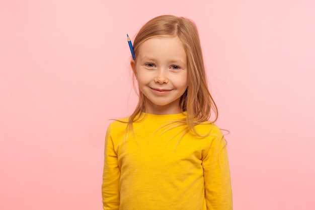 Retrato de encantadora menina adorável com cabelo ruivo, segurando o lápis atrás da orelha e sorrindo para o desenvolvimento da câmera de habilidades criativas da criança, tiro de estúdio interno isolado no fundo rosa