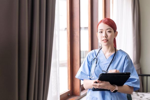 Retrato de enfermeira asiática usando estetoscópio de bata médica e segurando o tablet na casa do paciente Visita de cuidador em casa Cuidados de saúde em casa e conceito de lar de idosos