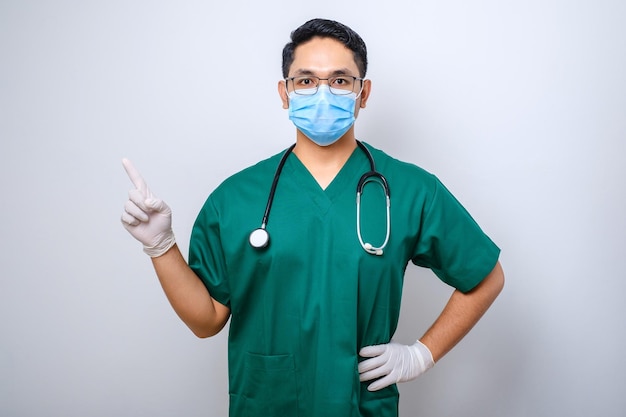 Retrato de enfermeiro masculino asiático apontando dedos para a esquerda usando máscara facial e luvas de borracha em uniforme clínico