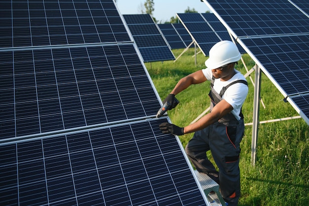 Retrato de engenheiro eletricista afro-americano em capacete de segurança e uniforme instalando painéis solares