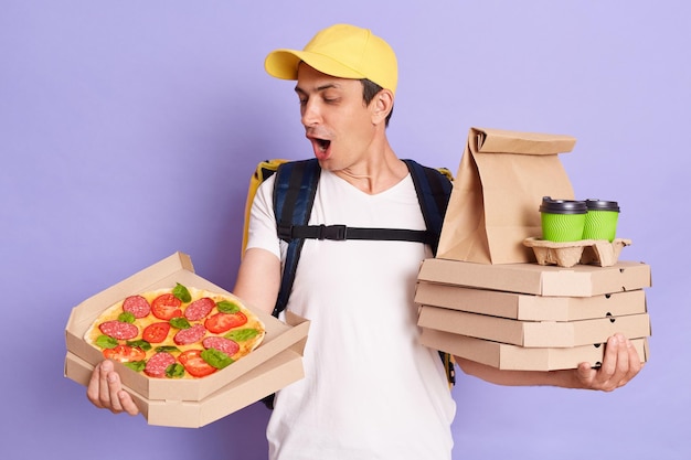 Retrato de entregador espantado usando boné amarelo segurando caixas de comida e café nas mãos olhando pizza saborosa com excitação posando isolado sobre fundo roxo