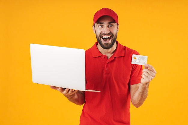 Retrato de entregador otimista de camiseta vermelha e boné segurando um laptop prateado e um cartão de crédito isolado sobre o amarelo