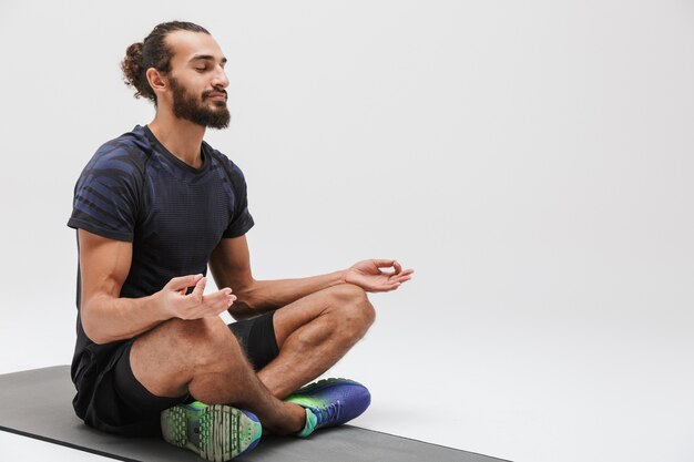 Retrato de esportista morena atlética com agasalho de treino meditando enquanto está sentado no tapete de ioga isolado no branco