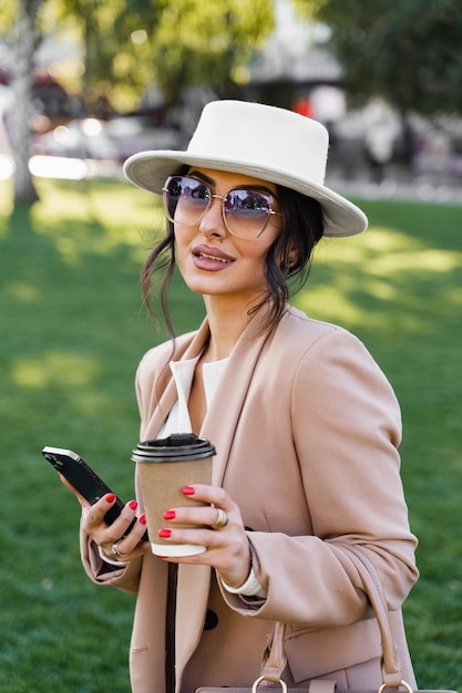 Retrato de estilo de rua do blogueiro com smartphone e xícara de café. garota usava chapéu branco, vestido e jaqueta.