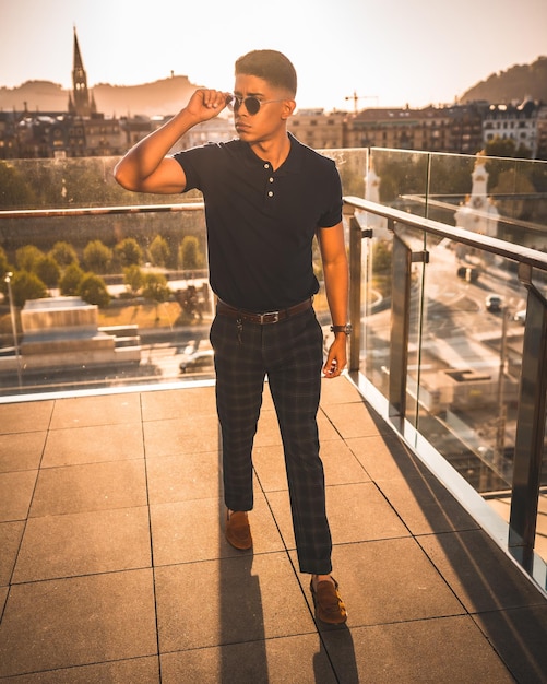 Retrato de estilo de vida de moda de um jovem latino com a cidade de San Sebastian ao fundo Gipuzkoa Camisa polo azul e calça xadrez passeando por do sol