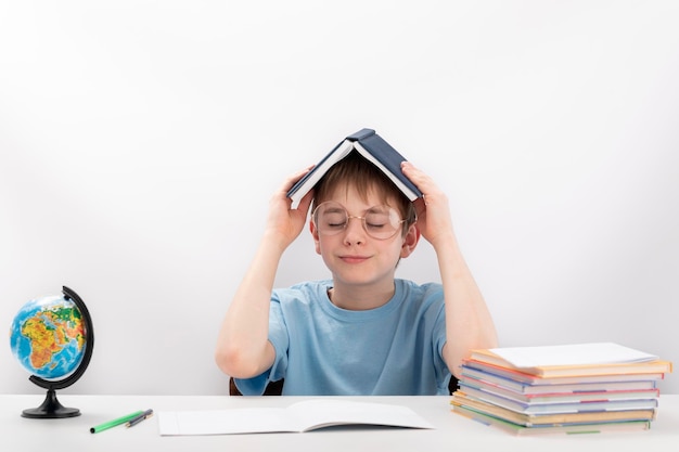 Retrato de estudante de óculos senta-se à mesa e segurando o livro sobre a cabeça Menino cansado com pilha de livros e cadernos escolares