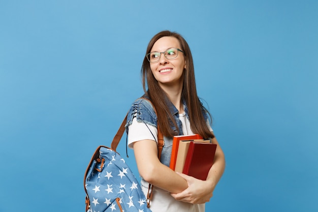 Retrato de estudante jovem casual bonita de óculos com mochila, olhando de lado no espaço da cópia, segurando o livro escolar isolado sobre fundo azul. Educação no conceito de faculdade de Universidade de ensino médio.