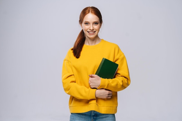 Retrato de estudante universitário alegre jovem segurando o livro e olhando para a câmera no fundo cinza isolado Modelo de senhora ruiva bonita vestindo roupas casuais emocionalmente mostrando expressões faciais