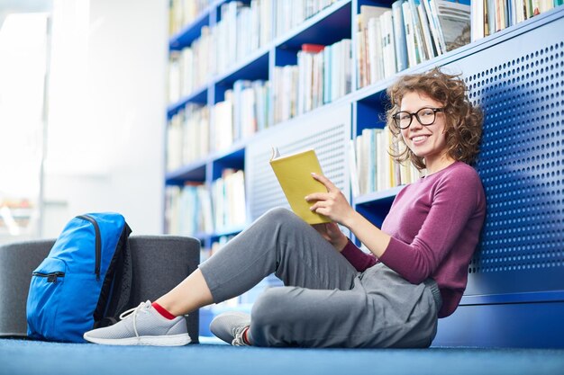 Foto retrato de estudante universitário alegre sentado no chão com um livro na biblioteca moderna