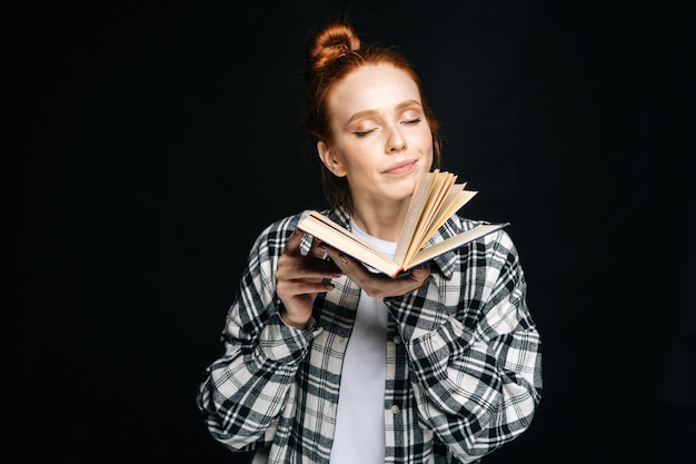 Retrato de estudante universitário jovem sorridente em pé com os olhos fechados, segurando o livro aberto no fundo preto isolado modelo de senhora ruiva bonita mostrando emocionalmente expressões faciais no estúdio