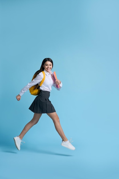 Retrato de estúdio completo de uma estudante adolescente caucasiana feliz em camisa casual branca e vestido preto carregando mochila amarela sorrindo olhando para a câmera correndo sobre fundo azul isolado