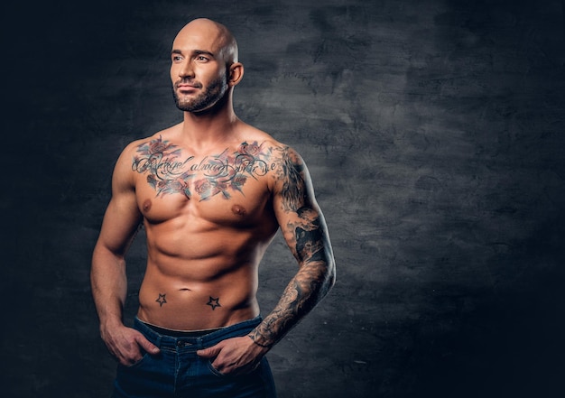 Retrato de estúdio de cabeça raspada sem camisa, macho musculoso com tatuagens no torso sobre fundo cinza vinheta.