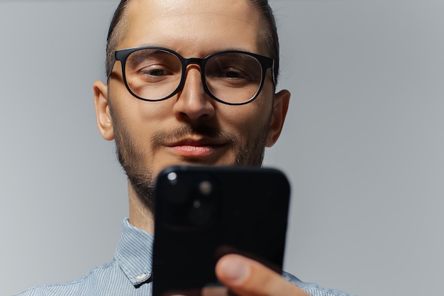 Retrato de estúdio de close-up de cara sorridente olhando no smartphone usando óculos em fundo cinza