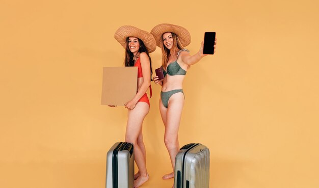 Retrato de estúdio de duas jovens mulheres em biquínis com chapéus de palha e malas prontas para sair de férias amigas sorridentes segurando um cartaz em branco um telefone celular e um passaporte com copyspace